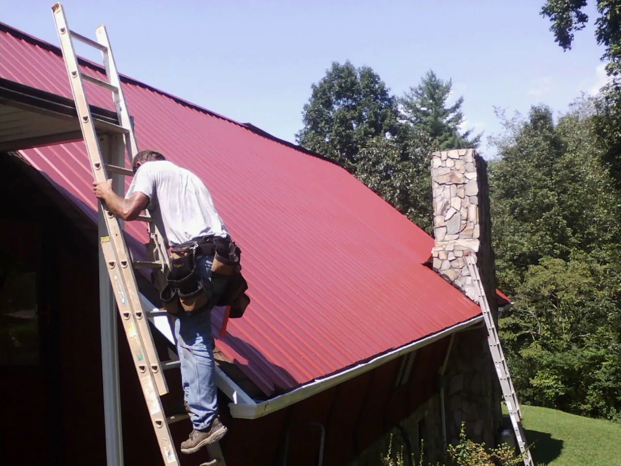 Roofer on a ladder beside a red metal roof and stone chimney — Soffit & Fascia Repair by Roofing Brothers in New Washington